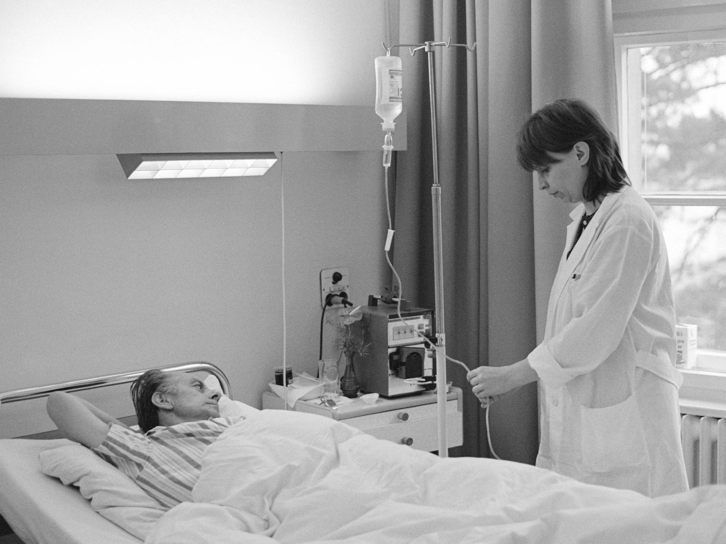 Fotografie, schwarz-weiß, von 1991: Das Foto zeigt einen, in einem Krankenhausbett liegenden Mann, der den Kopf nach rechts geneigt hat. Rechts steht eine Frau im weißem Kittel, die eine Infusion einstellt.